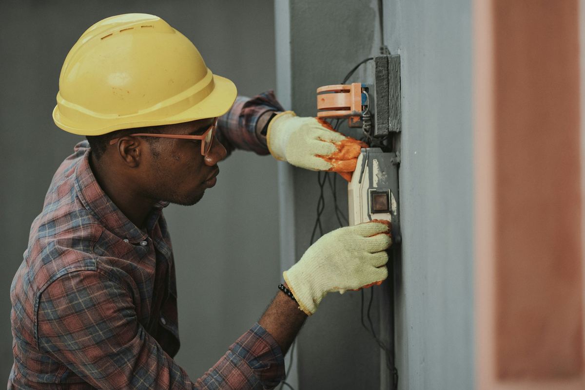 An electrician in a yellow hard hat and work gloves installs wiring on a job site
