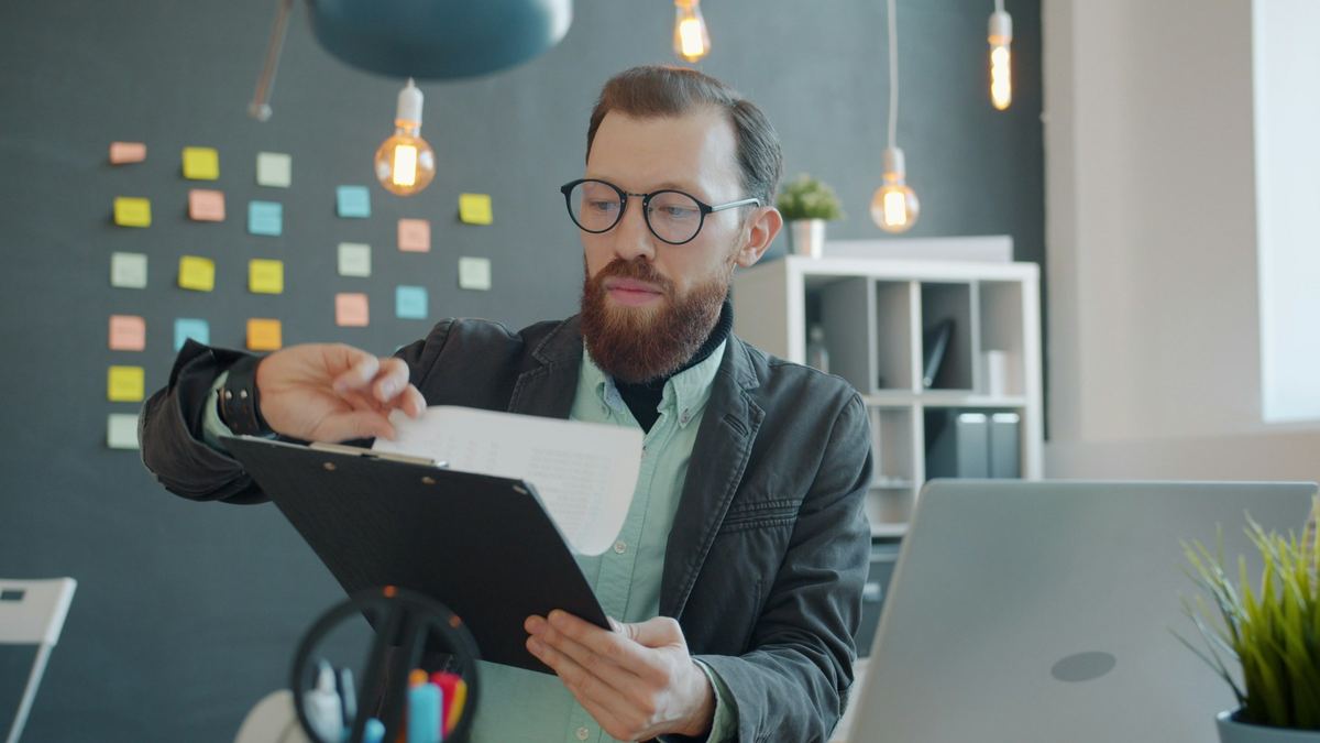 A man in glasses reads through printed reports at his desk, a laptop open beside him