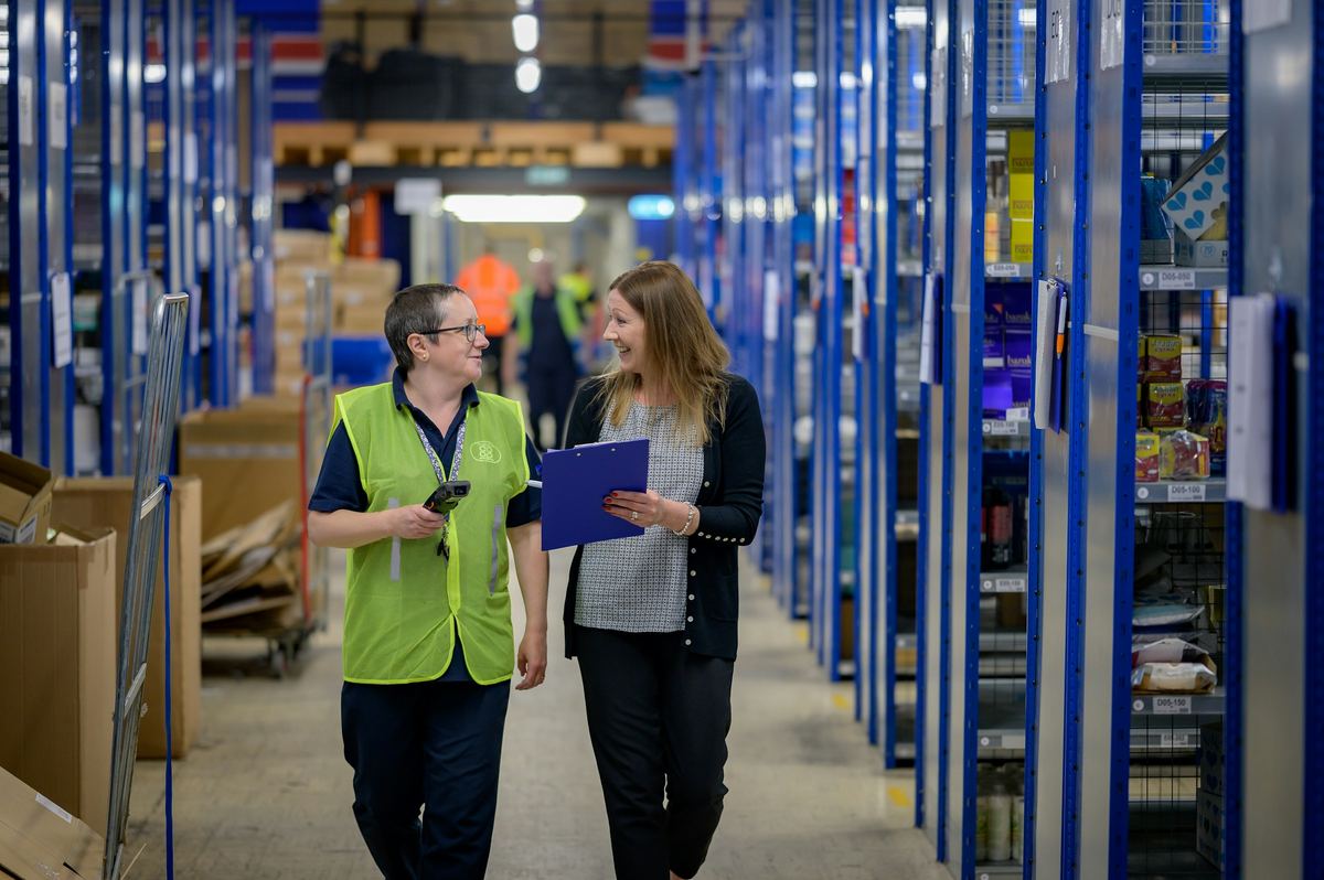 Two women walk a warehouse aisle lined with blue shelving, one carrying a clipboard