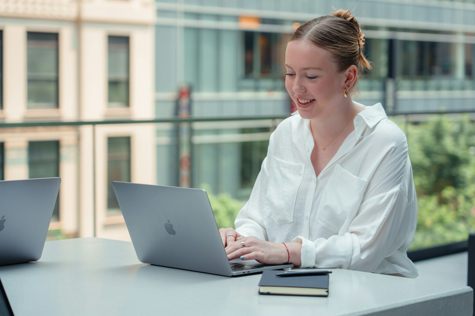 A smiling businesswoman in a white shirt works on her laptop at a bright office table, glass and greenery behind her